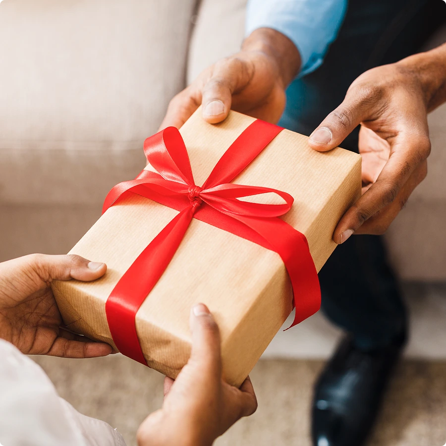 Close-up of two people exchanging a holiday gift wrapped in brown paper with a red ribbon.