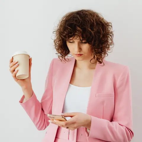 Woman in a pink suit holding a coffee while scrolling on a smartphone